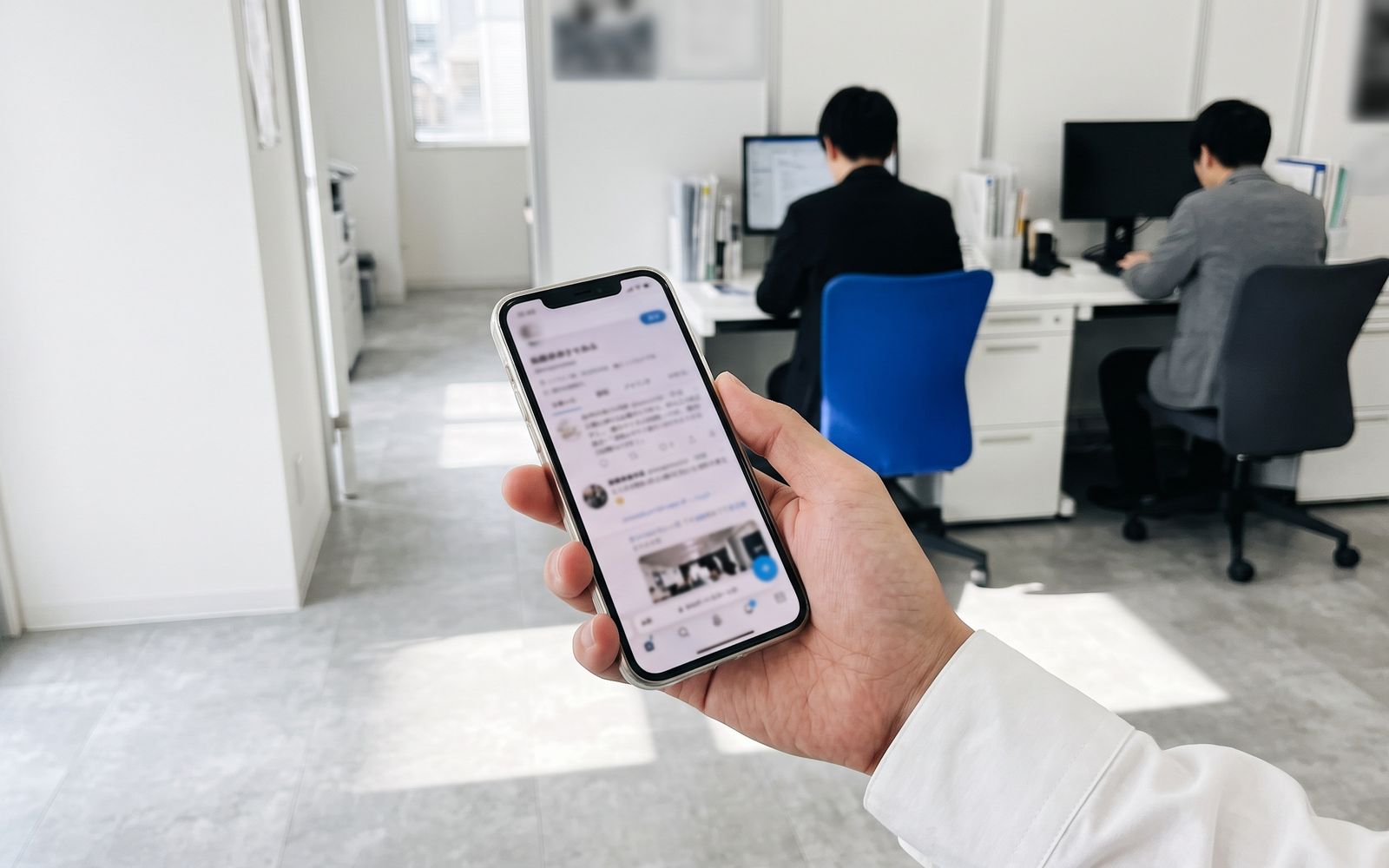 事務所の入口でスマートフォンの採用ページを見る手元と、奥で働く2人の後ろ姿
